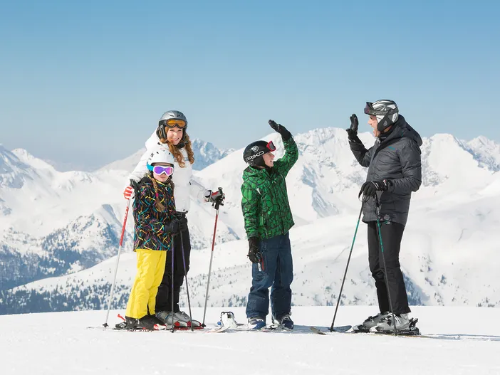 Familie in Skikleidung auf verschneitem Gipfel, Kinder klatschen ab, strahlend blauer Himmel